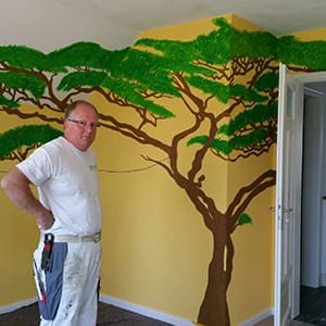 Thomas Huber vor einer gelben Wand mit gemaltem Baummotiv